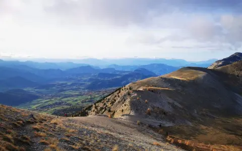 Un randonneur solitaire entame sa grande traversée des Alpes, dominant une vallée verdoyante sous un ciel nuageux.