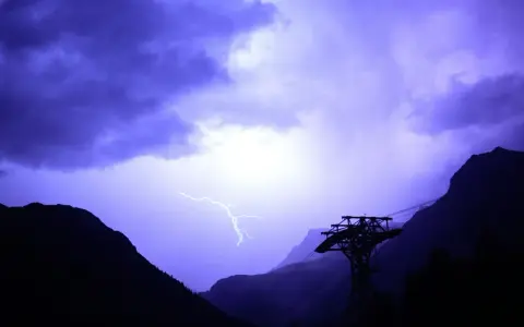 Un éclair zèbre le ciel violet d'un **orage en montagne que faire** ? Une cabine de téléphérique silhouette se dresse, témoin de la fureur de la nature.