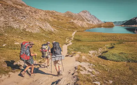 Trois randonneurs équipés pour le tour du Queyras traversent un sentier de montagne, un lac bleu et des sommets rocheux en toile de fond.