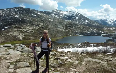 Jeune randonneuse souriante avec bâtons, face à une **mare** et des montagnes enneigées. La **difficulté** du sentier **nord** est palpable.
