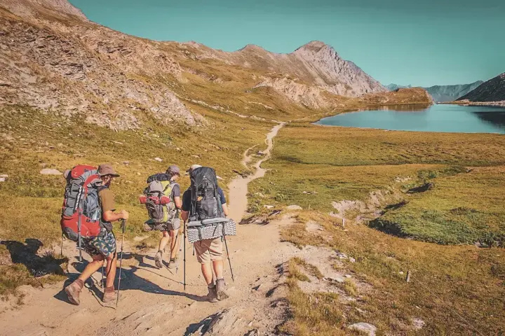 Trois randonneurs équipés pour le tour du Queyras traversent un sentier de montagne, un lac bleu et des sommets rocheux en toile de fond.