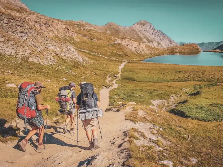 Trois randonneurs équipés pour le tour du Queyras traversent un sentier de montagne, un lac bleu et des sommets rocheux en toile de fond.