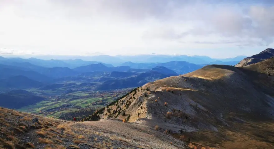 Un randonneur solitaire entame sa grande traversée des Alpes, dominant une vallée verdoyante sous un ciel nuageux.
