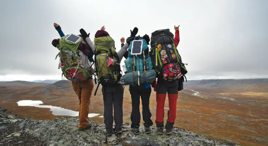 Quatre randonneurs, sacs à dos prêts, contemplent un paysage sauvage. Leur équipement, incluant des panneaux solaires, suggère une préparation pour une liste matériel randonnée 5 jours.
