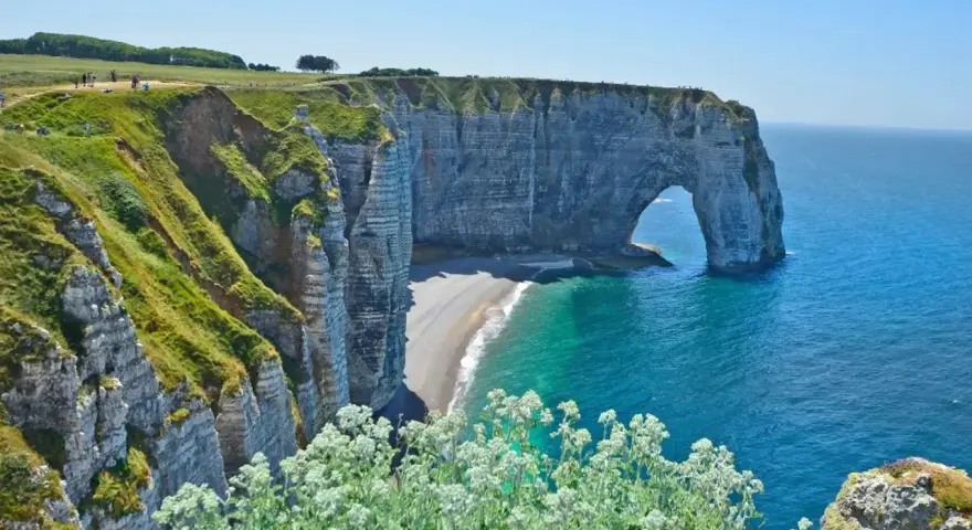 Falaises d'Étretat, France. L'arche naturelle de la Porte d'Aval se dresse fièrement au-dessus de la plage de galets. Le **gr 21** serpente le long de la côte verdoyante.