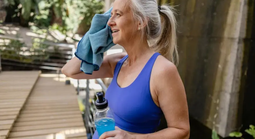 Femme d'âge mûr s'essuyant le visage avec une serviette bleue, tenant une bouteille d'eau. Elle fait du sport malgré la canicule.