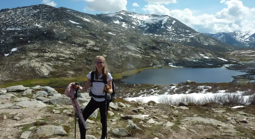 Jeune randonneuse souriante avec bâtons, face à une **mare** et des montagnes enneigées. La **difficulté** du sentier **nord** est palpable.