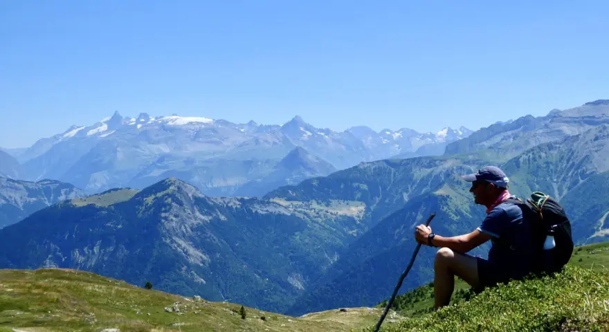 Un randonneur léger profite d'une vue panoramique sur les montagnes. Il est assis sur une colline verdoyante, bâton à la main, admirant le paysage majestueux.