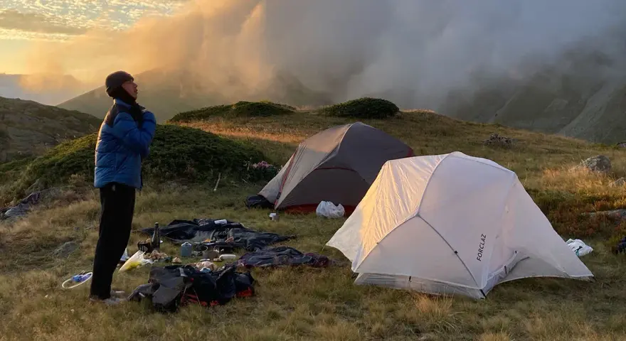 Une personne se tient près de tentes dans une aire de bivouac, profitant du lever du soleil sur les montagnes.