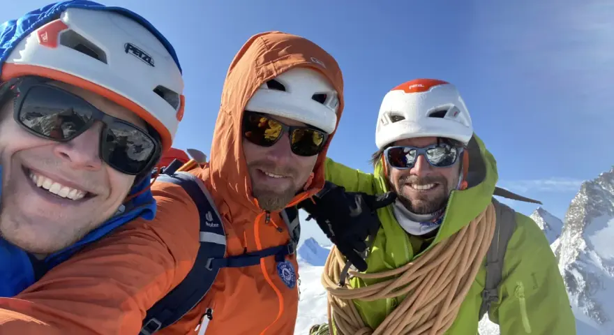 Trois alpinistes souriants au sommet, prêts à devenir guide de haute montagne à 40 ans. Corde et équipement de sécurité visibles.
