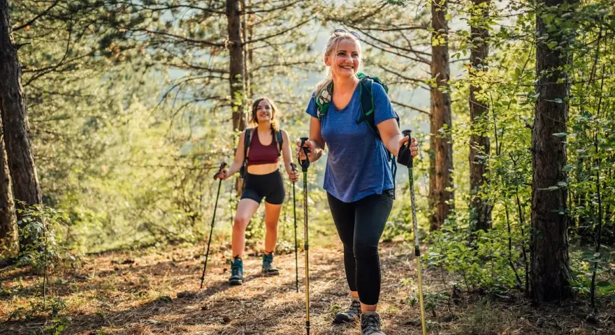 Deux femmes souriantes randonnent en forêt, utilisant des bâtons. Elles semblent apprécier la nature, un argument pour ou contre les bâtons de randonnée.