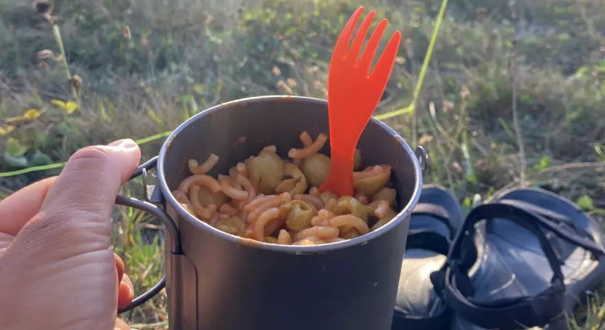 Un repas simple pour savoir que manger en randonnée : des pâtes avec des olives dans une tasse de camping, avec une fourchette orange.