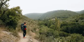 Randonneuse avec sac à dos sur un chemin d'amadou, contemplant une vallée verdoyante. La carte du chemin d'amadou est dans sa poche.
