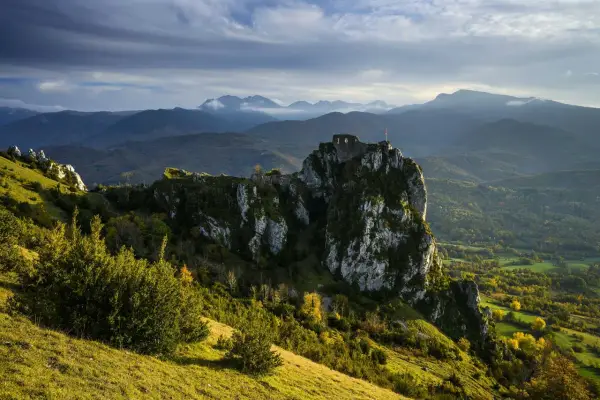 Ruines d'un château cathare sur un pic rocheux, dominant un paysage de montagnes et de forêts. Le chemin des bonshommes serpente à travers la vallée.