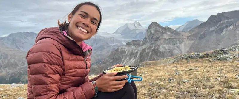 Sourire radieux d'une randonneuse savourant son repas en pleine montagne, avec des sommets enneigés en arrière-plan. Un moment de pur bonheur pour ce repas randonnée.