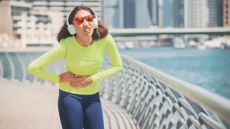 Une coureuse en tenue de sport, écouteurs aux oreilles, se tient le ventre, visiblement prise d'un point de côté.
